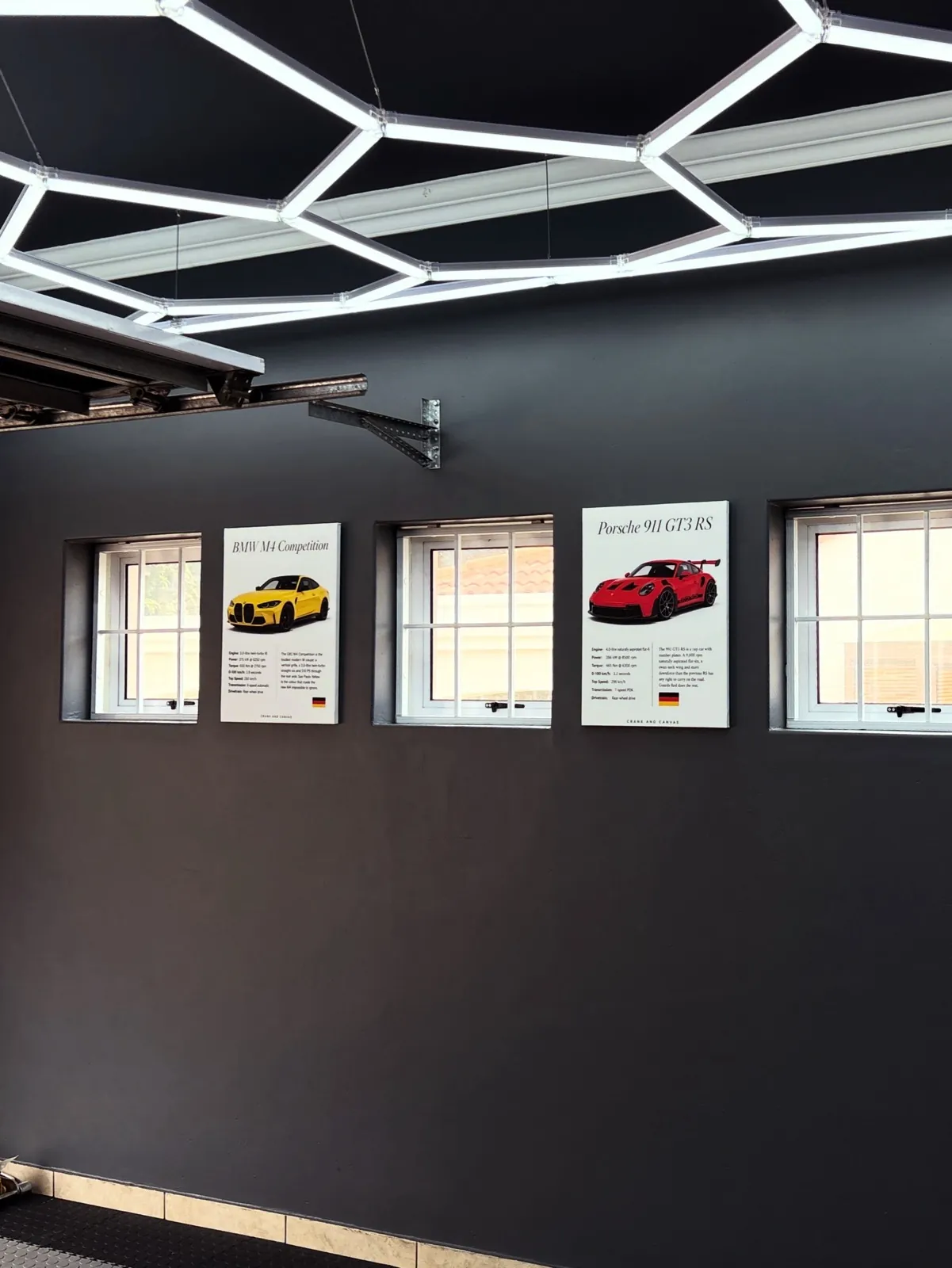 BMW M4 Competition and Porsche 911 GT3 RS canvases on a charcoal garage wall under hexagon lighting